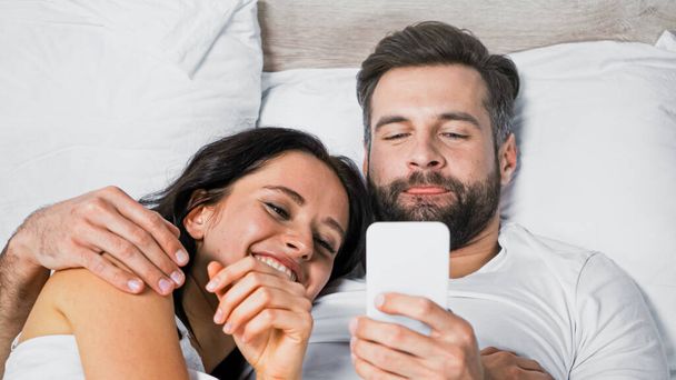 man hugging cheerful girlfriend while using smartphone in bed - Фото, изображение