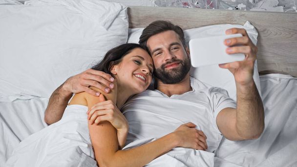 szczęśliwy brodaty mężczyzna obejmujący wesoły dziewczyna podczas biorąc selfie w łóżku - Zdjęcie, obraz