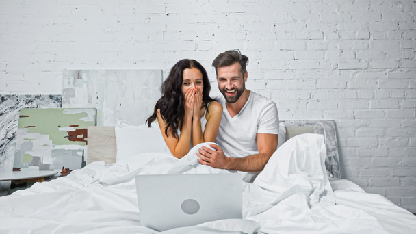 excited woman covering mouth with hands while watching comedy on laptop with laughing boyfriend - Photo, Image