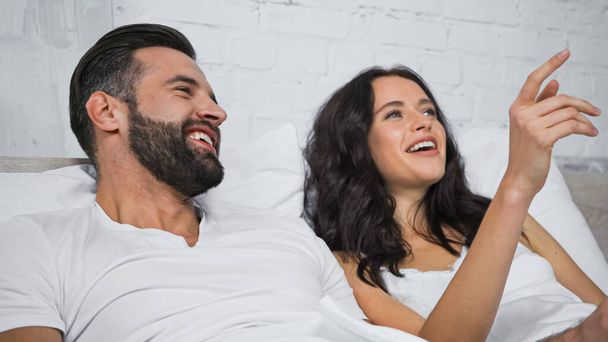 excited brunette woman looking away and pointing with finger near cheerful boyfriend in bed - Photo, Image