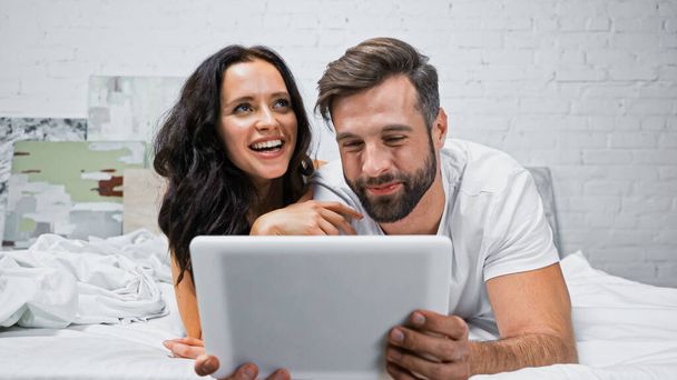 mujer emocionada riendo cerca de novio sonriente con tableta digital en el dormitorio - Foto, imagen
