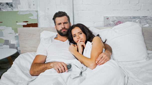 tense man embracing worried woman while watching tv in bedroom - Foto, afbeelding