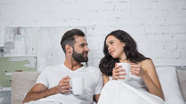 happy young couple with cups of coffee talking in bed - Фото, изображение