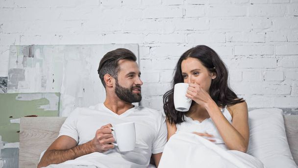 happy bearded man looking at girlfriend drinking coffee in bed - Photo, image