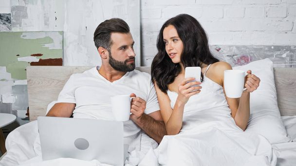 jeune couple avec des gadgets tenant des tasses et se regardant dans le lit - Photo, image