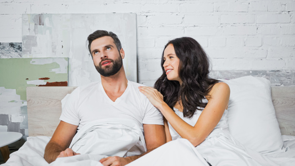 cheerful woman touching shoulder of bored boyfriend while talking to him in bed - Valokuva, kuva