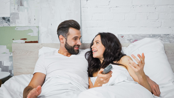 cheerful couple looking at each other while watching tv in bed - Photo, Image