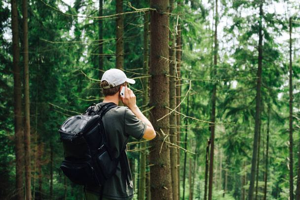 Günlük giysiler içinde bir erkek turist ve sırtında bir sırt çantası dağlardaki bir akıllı telefondan iletişim kuruyor, ormanda duruyor ve çağrının üzerine dinleniyor.. - Fotoğraf, Görsel