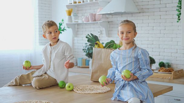 Lächelndes Mädchen mit Äpfeln auf Küchentisch neben Bruder und Papiertüte  - Foto, Bild