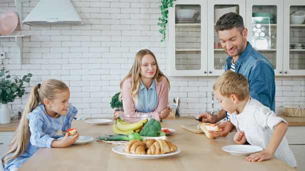 Улыбающийся мужчина дает багет сыну рядом с семьей на кухне  - Фото, изображение