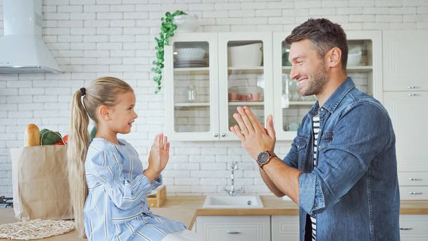 Side view of smiling father and daughter playing patty cake game in kitchen  - Foto, Bild