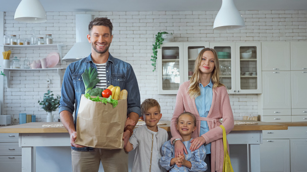 Lächelnder Mann hält Papiertüte mit frischen Lebensmitteln in der Nähe einer Familie mit Kindern in der Küche  - Foto, Bild