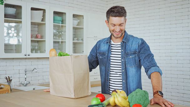 Smiling man looking at fresh food near paper package on kitchen table  - Φωτογραφία, εικόνα