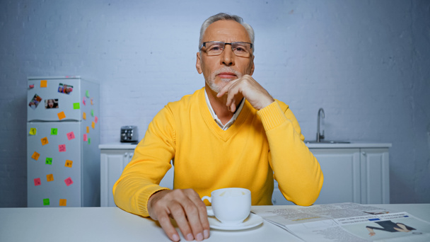 Idősebb férfi szemüvegben a kamerát nézi egy csésze kávé és újság közelében elmosódott előtérben  - Fotó, kép