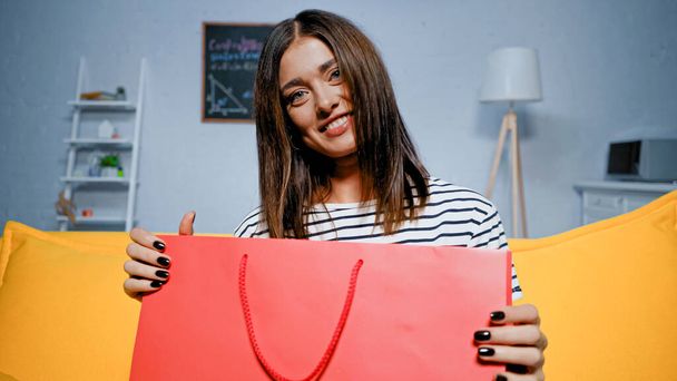 Młoda kobieta uśmiecha się do kamery trzymając torbę na zakupy w domu  - Zdjęcie, obraz