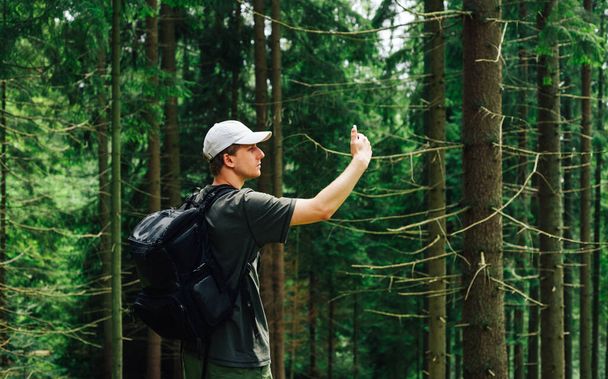 Handsome young male hiker on a hike stands in the woods with a smartphone in his hand and takes a photo of a beautiful natural landscape in the mountains. - Fotoğraf, Görsel