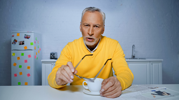 Elderly man holding eyeglasses while talking during video chat at camera in kitchen  - Φωτογραφία, εικόνα