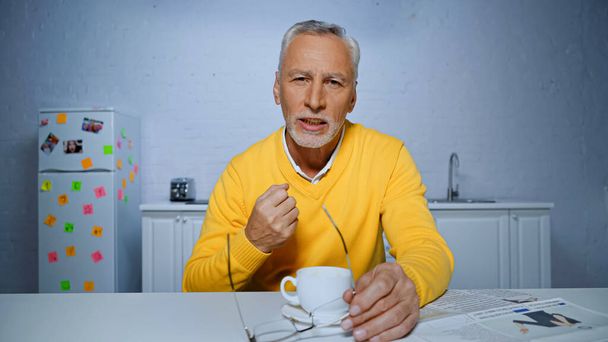 Elderly man holding eyeglasses and showing yeah gesture near cup during video call  - Фото, зображення