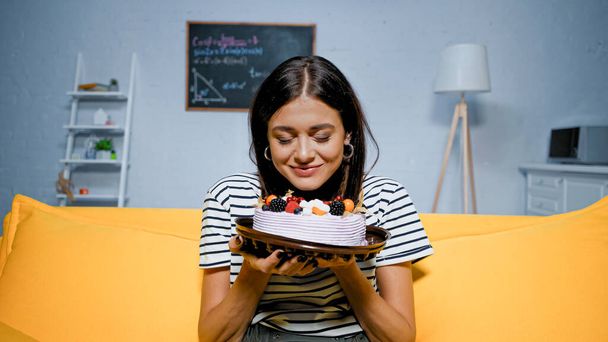 Mujer joven sosteniendo sabroso pastel con los ojos cerrados en la sala de estar  - Foto, Imagen
