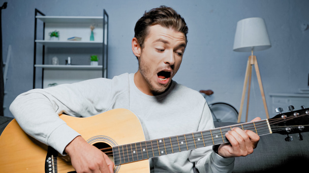 Νεαρός που παίζει ακουστική κιθάρα και τραγουδάει στο σπίτι  - Φωτογραφία, εικόνα