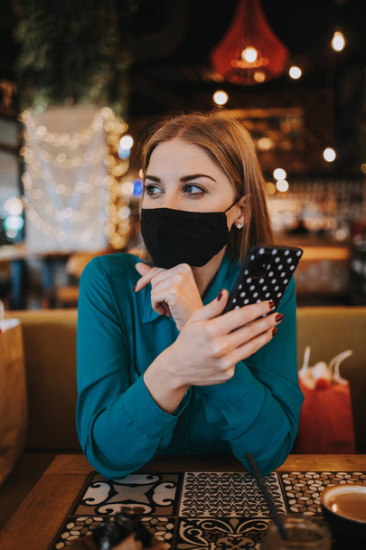 Beautiful young adult woman with protective face mask sitting in a nice restaurant and using her smart phone. She is alone and serious. Coronavirus pandemic and social distancing problems.  - Foto, immagini