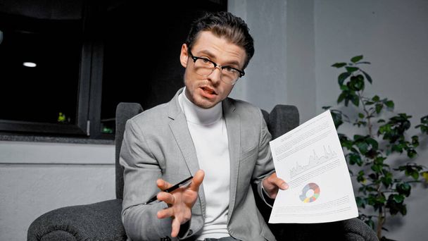 Joven hombre de negocios hablando y sosteniendo documento durante la videollamada  - Foto, imagen