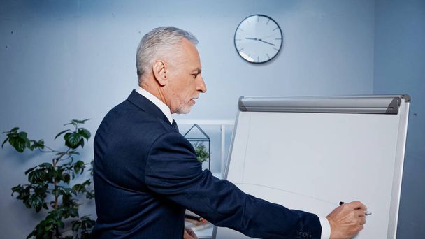 Älterer Geschäftsmann schreibt mit Filzstift auf Flipchart im Büro  - Foto, Bild