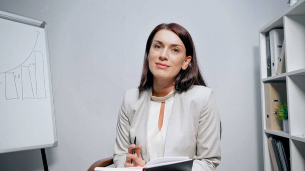 Geschäftsfrau mit Stift und Copybook blickt im Büro in die Kamera  - Foto, Bild