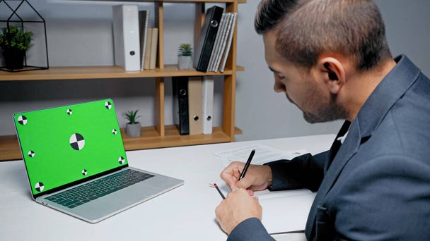 Laptop mit grünem Bildschirm neben Geschäftsmann, der in Notizbuch auf verschwommenem Vordergrund schreibt  - Foto, Bild