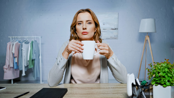 Žena drží pohár a dívá se do kamery v blízkosti notebooku na stole  - Fotografie, Obrázek