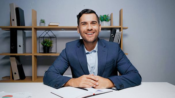 Smiling businessman looking at camera near notebook and papers on blurred foreground  - Photo, Image