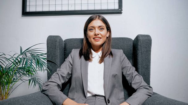 Smiling businesswoman sitting in armchair near plant in office  - Foto, afbeelding
