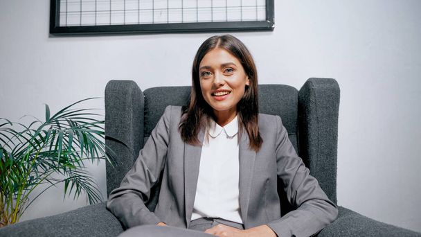 Young brunette businesswoman looking at camera on armchair in office  - Photo, Image