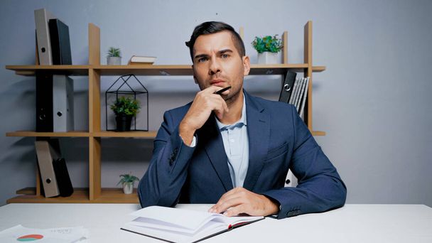 Pensive бізнесмен з ручкою дивиться на камеру біля ноутбука
  - Фото, зображення