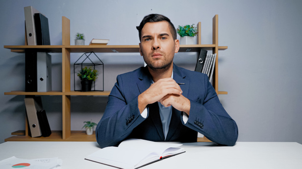 Бизнесмен смотрит в камеру возле ноутбука на столе  - Фото, изображение