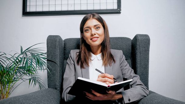 Smiling businesswoman holding notebook and pen in office  - Фото, изображение