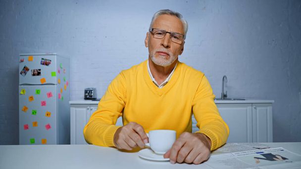 Senior man looking at camera near newspaper and coffee cup on blurred foreground  - Foto, Imagen
