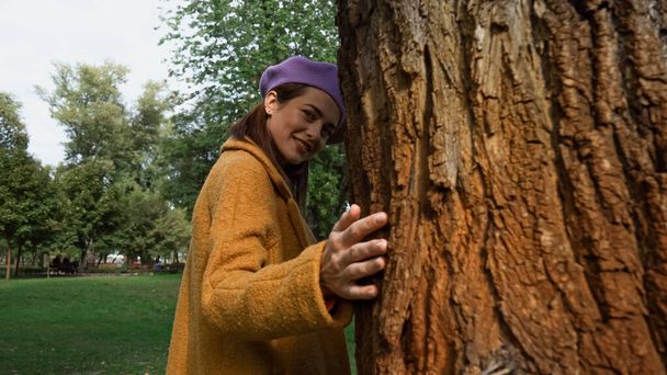 Mutlu kadın parktaki ağaç gövdesine dokunurken kameraya bakıyor. - Fotoğraf, Görsel