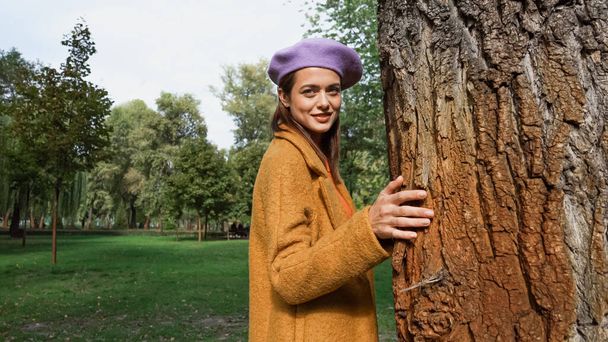 young fashionable woman looking at camera while standing near tree trunk in park - Foto, Imagem