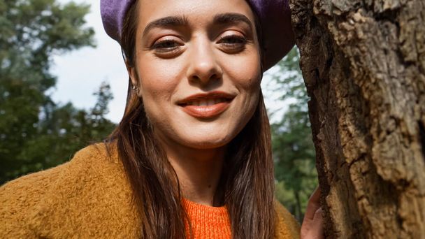 portrait of cheerful woman smiling at camera near tree trunk in autumn park - Photo, Image