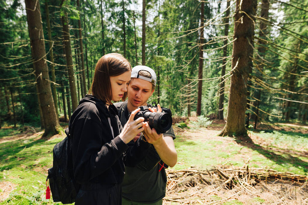 Man en vrouw staan in een zonnig bergwoud en bekijken foto 's vanaf het camerascherm en communiceren. Een paar wandelaars maken prachtige foto 's op een klim de berg op. Toerisme. - Foto, afbeelding