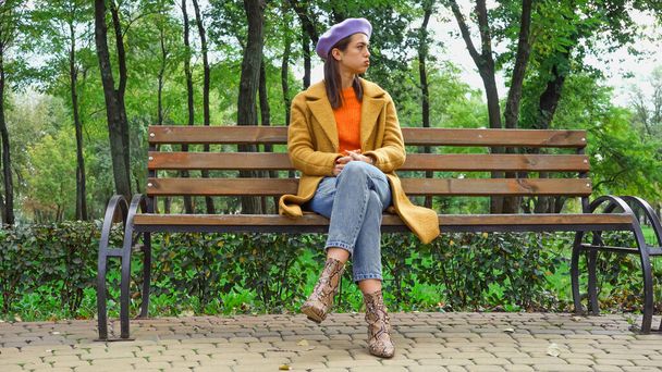 displeased stylish woman sitting on bench in park and looking away - Fotoğraf, Görsel