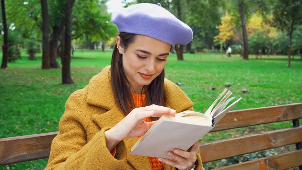 Улыбающаяся женщина в модном осеннем наряде читает книгу в парке - Фото, изображение