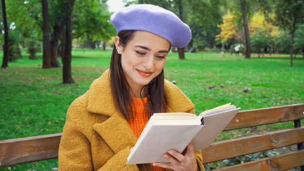 jonge stijlvolle vrouw glimlachen tijdens het lezen van roman in de herfst park - Foto, afbeelding