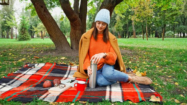 woman in stylish autumn clothes opening thermos during picnic in park - Valokuva, kuva