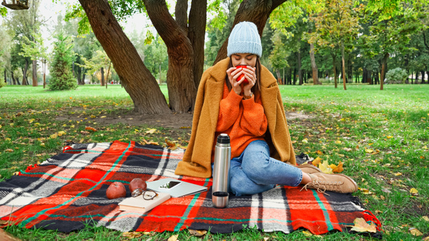 Frau im Herbst-Outfit trinkt warmen Tee auf karierter Decke im Park - Foto, Bild