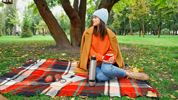 smiling woman in autumn outfit looking away while sitting on checkered blanket in park - 写真・画像