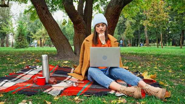 freelancer pensativo olhando para laptop enquanto sentado em cobertor xadrez no parque - Foto, Imagem