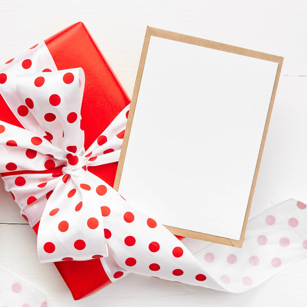 Greeting card mockup and red gift box with big bow on white wooden background, top view, copy space - Fotó, kép