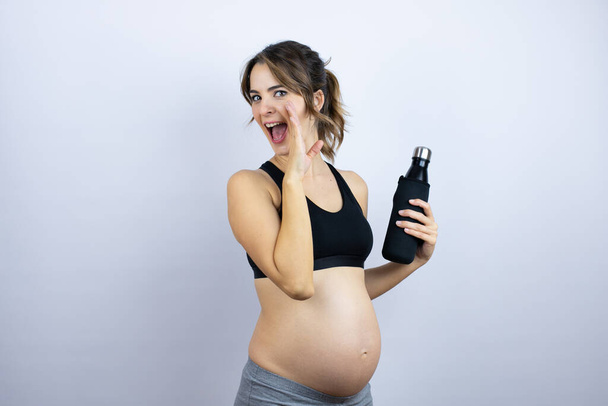 Mladá sportovkyně těhotná nosí sportovní oblečení drží láhev s vodou na bílém pozadí ruku na ústech vyprávění tajné zvěsti, šeptání škodolibý rozhovor - Fotografie, Obrázek